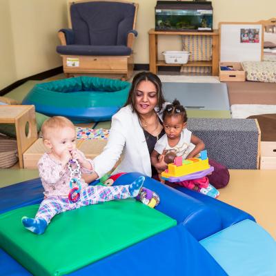 A woman is holding a baby sitting on the floor and reaching out to another baby sitting on a beanbag chair. 